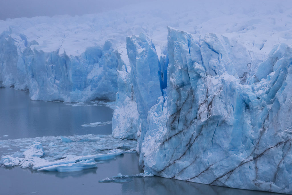 Sous la brume se dévoile lentement le célèbre glacier de Patagonie, un spectacle à la fois visuel et sonore : craquements, fissures et blocs de glace tombant avec fracas dans les eaux bleues du lac Argentino.